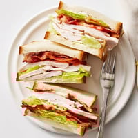 Golden toasted triple-decker Turkey Club Sandwich with crispy bacon, fresh lettuce, and tomato on a plate, served with potato chips.